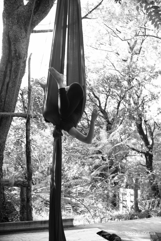 Amaya Lachaize-Müller enroulée dans un tissu aérien sombre, en extérieur parmi les arbres. Photographie en noir et blanc.
